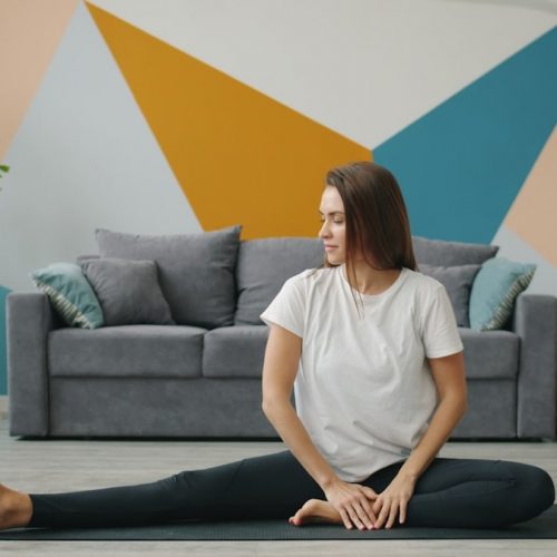 Attractive girl in leggings and T-shirt is doing stretching exercises on yoga mat relaxing on floor at home busy with practise. Youth, lifestyle and health concept.