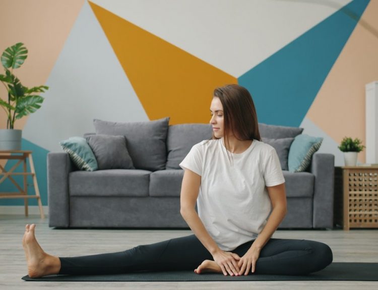 Attractive girl in leggings and T-shirt is doing stretching exercises on yoga mat relaxing on floor at home busy with practise. Youth, lifestyle and health concept.